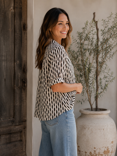 A woman wearing a black and white patterned short-sleeve blouse and light blue jeans, standing in a rustic indoor setting next to a large wooden door and a potted plant.