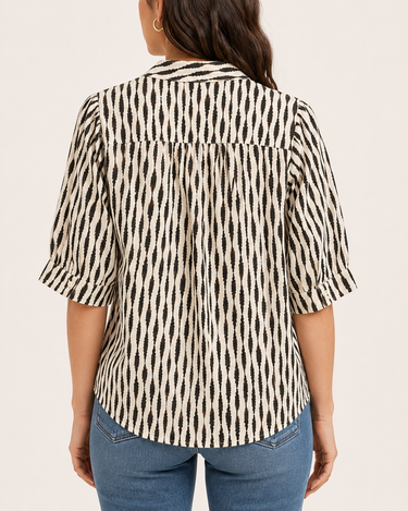 A woman viewed from the back wearing a short-sleeve button-up blouse with a black and cream ikat-style vertical pattern, paired with blue jeans, against a solid light beige background.