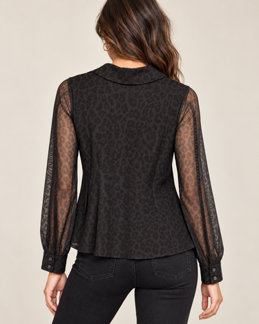A woman shown from the back wearing a black leopard-print long-sleeve blouse with sheer mesh sleeves and black jeans, against a plain light beige studio background.