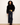 A model stands against a plain off-white wall wearing a black velvet jacket over a black top, dark wide-leg jeans, and brown leather boots, with a yellow plaid shirt tucked into the jeans.