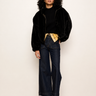 A model stands against a plain off-white wall wearing a black velvet jacket over a black top, dark wide-leg jeans, and brown leather boots, with a yellow plaid shirt tucked into the jeans.