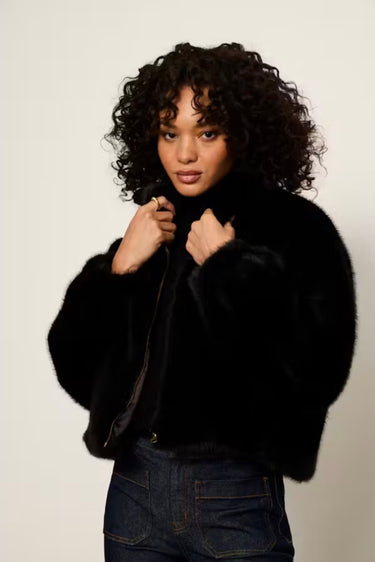 A model with curly dark hair wearing a black cropped faux fur jacket over a black top and dark blue jeans, posing against a light gray studio background.