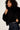 A model with curly dark hair wearing a black fuzzy faux-fur jacket over a black top and dark blue jeans, posing against a plain off-white background.