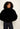 A model with curly black hair wearing a black faux fur cropped jacket and dark blue jeans, standing against a plain off-white background.