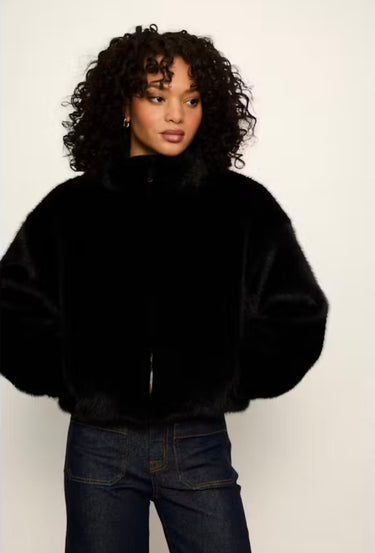 A model with curly black hair wearing a black faux fur cropped jacket and dark blue jeans, standing against a plain off-white background.