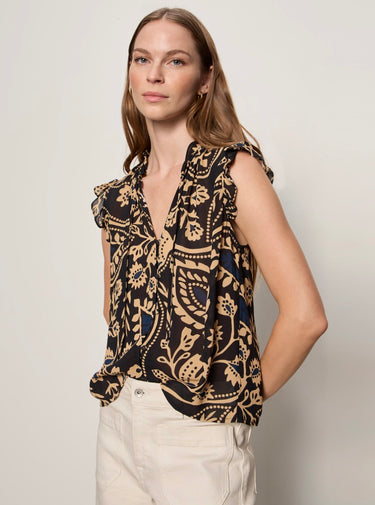 A woman wearing a sleeveless black blouse with a beige and navy blue paisley pattern, paired with cream-colored pants, photographed against a neutral light gray studio background.