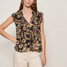 A woman shown from the back wearing a sleeveless black blouse with a beige paisley pattern and ruffled shoulders, paired with cream pants, against a light gray studio background.