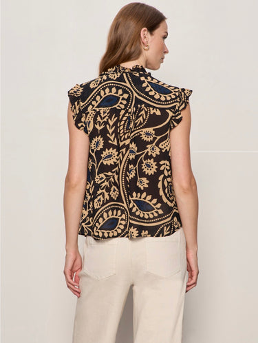 A woman shown from the back wearing a sleeveless black blouse with a beige paisley pattern and ruffled shoulders, paired with cream pants, against a light gray studio background.