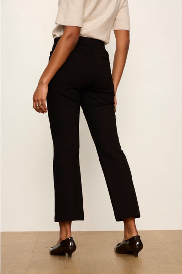 A model shown from the back wearing black cropped ankle-length trousers, a cream short-sleeve top, and black patent leather kitten heels against a plain off-white wall and light wood floor.
