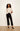 A woman modeling a cream short-sleeve knit top and black cropped trousers with black pointed-toe heels, standing against a plain off-white wall.