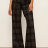 A model wearing high-waisted, wide-leg plaid trousers in a dark brown and black tartan pattern, paired with black leather shoes, standing against a plain off-white wall.