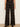 A model is shown from the waist down wearing high-waisted, wide-leg plaid trousers in a dark brown and black pattern, paired with black pointed-toe heels against a neutral cream background.