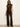 A model wearing a brown satin mock-neck top and dark brown/black plaid flared trousers, standing against a plain off-white wall.