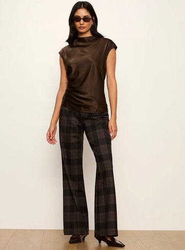 A model wearing a brown satin mock-neck top and dark brown/black plaid flared trousers, standing against a plain off-white wall.