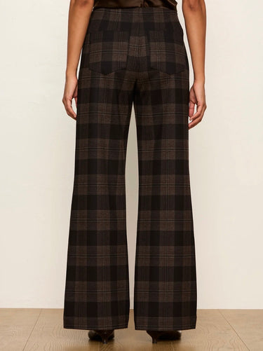 A person shown from the back wearing dark brown and black plaid wide-leg trousers, standing against a plain off-white wall on a light wood floor.