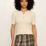 A woman with curly black hair wearing a cream short-sleeve henley top and olive green plaid pants, standing against a plain off-white background.