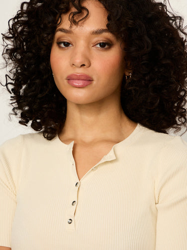 A close-up shot of a woman with curly dark hair wearing a cream-colored ribbed short-sleeve henley top with a buttoned collar.