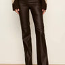 A person wearing dark brown faux-leather flare pants with a ribbed knit sweater and black boots, standing against a plain off-white wall.