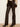 A model is shown from the waist down wearing dark brown faux-leather flare pants, a beige and brown knit sweater, and black pointed-toe heeled boots against a neutral cream wall and wood floor.