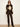 A woman modeling a dark brown and beige argyle sweater with wide sleeves, paired with dark brown leather pants and black pointed-toe heels, against a plain off-white studio background.