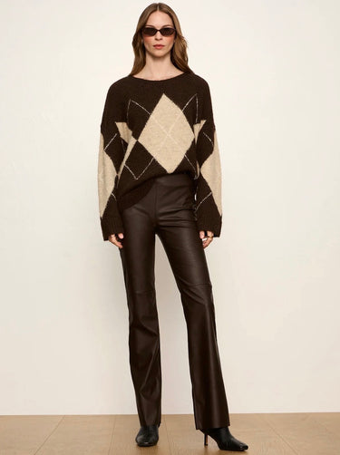 A woman modeling a dark brown and beige argyle sweater with wide sleeves, paired with dark brown leather pants and black pointed-toe heels, against a plain off-white studio background.