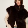 A woman wearing a dark brown fur vest over a cream lace long-sleeve top and blue jeans, photographed against a plain light background.