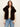 A woman modeling a dark brown faux fur vest over a cream lace long-sleeve top and blue jeans, photographed against a plain off-white studio background.