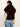 A woman shown from the back wearing a dark brown fur vest over a cream lace long-sleeve top and blue jeans, against a plain off-white studio background.