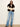 A model stands against a plain off-white wall wearing a dark brown fur vest over a cream lace long-sleeve top, paired with light-wash blue jeans and black pointed shoes.
