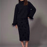 a woman wearing a black sweater with feather cuffs, a mesh midi skirt with a velvet floral pattern and black heels on a grey studio background