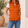 a woman wearing a shiny pedant puffer vest, a white tee, blue jeans, and white sneakers on a light grey studio backdrop.