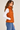 a woman facing sideways wearing a shiny pedant puffer vest, a white tee, blue jeans, and white sneakers on a light grey studio backdrop.