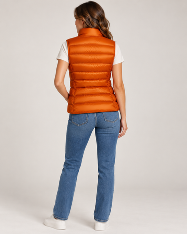 a woman facing backwards wearing a shiny pedant puffer vest, a white tee, blue jeans, and white sneakers on a light grey studio backdrop.