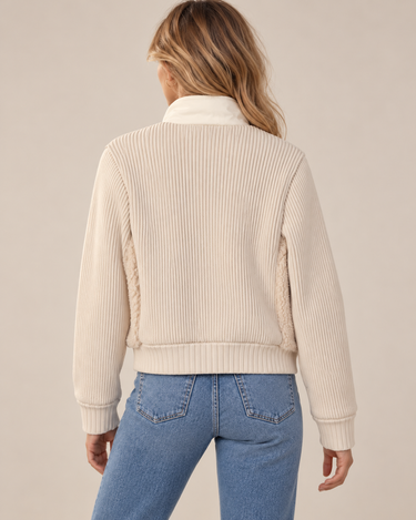 A woman wearing a ribbed beige jacket and blue jeans on a neutral background.