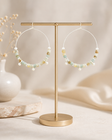 A pair of large hoop earrings with small pastel beads and pearl-like accents, displayed on a gold-tone stand against a soft neutral background.