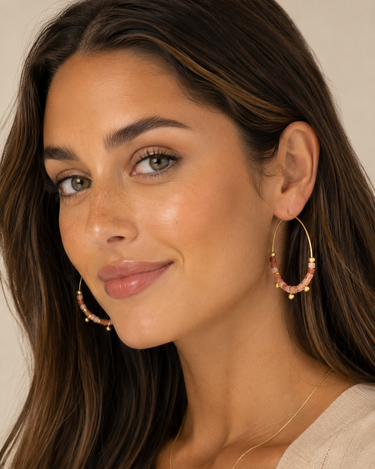 A close-up of a woman wearing large gold hoop earrings adorned with pink and white beads, set against a soft beige background.