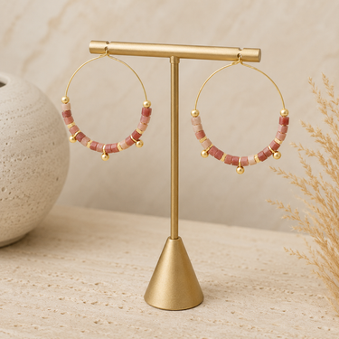 Gold-tone hoop earrings with pink and cream beaded details, displayed on a gold T-stand against a neutral beige background.
