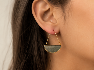 A close-up of a model's ear wearing a gold hoop earring with a semi-circular green stone pendant.