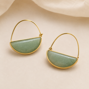A pair of gold hoop earrings with a polished, semi-circular light green stone inlay, displayed on a soft beige fabric background.