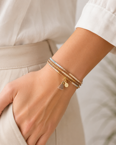 A close-up of a person's wrist wearing a stack of delicate bracelets, including a gold chain with a small gold tassel charm and a silver-toned beaded bracelet, paired with a white blouse and beige trousers.