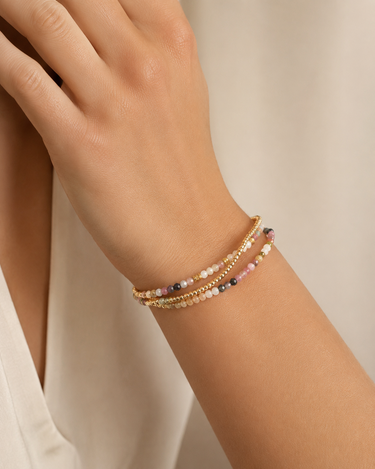 A close-up of a person's wrist wearing three delicate beaded bracelets, including a gold chain and strands of small multicolored beads, against a soft cream-colored background.