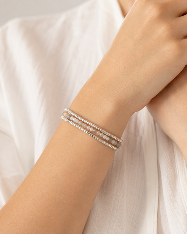 A close-up of a person's wrist wearing a delicate silver double-strand bracelet with small clear stones and a central row of white pearls, set against a soft white draped fabric background.