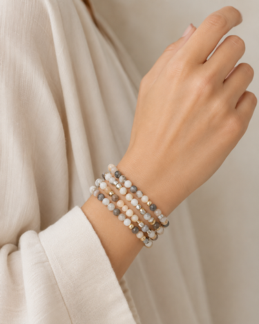 A close-up of a person's wrist wearing three beaded stretch bracelets in neutral tones of beige, gray, and white, styled with a cream-colored top.