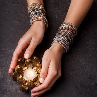 Hands holding a decorative candle holder with a lit candle, wearing beaded bracelets on a dark background.