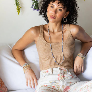 Woman wearing a beige tank top and floral pants, sitting on a white couch with a neutral background.