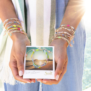 Person holding a card with a bracelet design, wearing multiple colorful bracelets on a blurred natural background