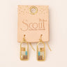 A pair of gold-tone dangle earrings with rectangular wood inlay panels in natural, beige, and light blue tones, displayed on a peach-colored card labeled Scout Curated Wears.