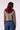 A woman shown from the back wearing a cropped, long-sleeve blazer-style top with a brown and beige houndstooth pattern and a contrasting dark red hood, paired with blue jeans, against a light studio background.