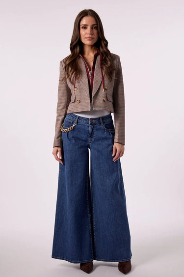 A woman modeling a cropped brown plaid blazer with gold buttons, a red collar detail, and a gold chain accessory, paired with wide-leg blue jeans and brown boots, against a light gray studio background.