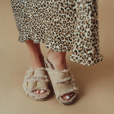 A person wearing beige fuzzy slide sandals with gold-tone buckle straps, shown from the knees down against a warm beige studio background. A leopard-print skirt is also visible.